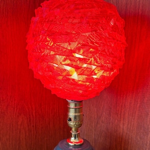May include: A table lamp featuring a red, textured lampshade. The shade is supported by a brass neck and a teal blue ceramic base. The lamp is lit, emitting a warm light. The background is a reddish-brown wooden surface.