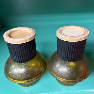 May include: Two vintage salt and pepper shakers. The shakers have a gold-colored, bulbous base, a black, ribbed midsection, and a light-colored top. The shakers are displayed on a turquoise surface.