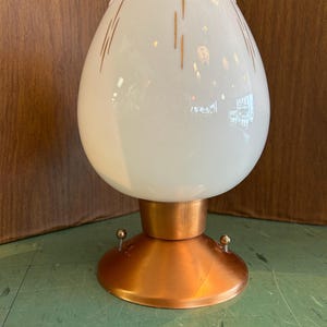 May include: A vintage table lamp featuring a white, egg-shaped glass shade with vertical gold lines. The shade rests on a copper-coloured base with a round, flared design and small, decorative brass accents. This is a retro lighting fixture.