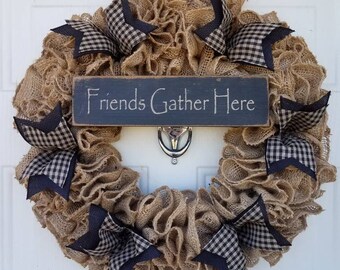 May include: A rustic burlap wreath with a sign that reads "Friends Gather Here." The wreath features a ruffled texture and is adorned with navy blue and black gingham star-shaped bows. A decorative piece for the front door.