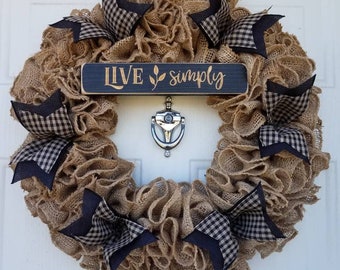 May include: A rustic burlap wreath with navy blue and black gingham bows. A small, dark blue wooden sign in the center reads "LIVE simply" in gold lettering. A silver door knocker is below the sign.