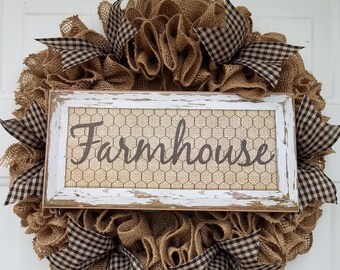May include: A farmhouse-style wreath with a burlap base and a white distressed wooden sign. The sign features the word "Farmhouse" in a script font over a chicken wire background. Black and white checkered ribbons are incorporated.