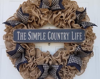 May include: A rustic burlap wreath with a sign that reads "The Simple Country Life." The wreath is adorned with navy blue and black checkered bows. The wreath is a decorative item for a home.