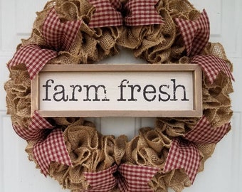 May include: A rustic burlap wreath with red and white checkered ribbon accents. The wreath features a wooden sign in the center that reads "farm fresh" in black lettering. This farmhouse-style wreath is perfect for a front door or interior decor.