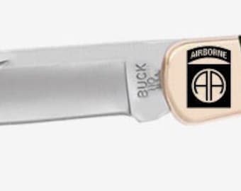 May include: A Buck 110 folding knife with a black wood handle and a gold-coloured blade. The blade has an inscription that reads "Airborne" and a symbol of a parachute with the letters "AA" inside. The handle has a gold-coloured inscription that reads "J.F."