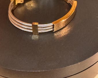 May include: A gold and silver bracelet with a clasp. The bracelet features three silver-colored, woven bands and gold-colored rectangular accents. The bracelet is displayed on a black surface.