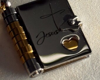 May include: A silver-colored pendant necklace in the shape of a small book. The book has a cross and the word "Jesus" engraved on the cover. A heart-shaped cutout reveals a gold-colored interior. The pendant is attached to a cylindrical container with gold and silver accents.