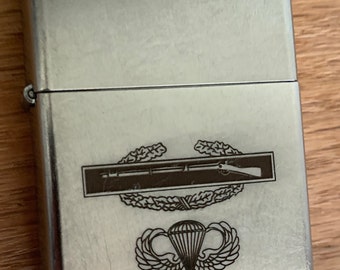 May include: Silver lighter with an engraved design of a military insignia. The insignia features a rifle and a parachute with wings.