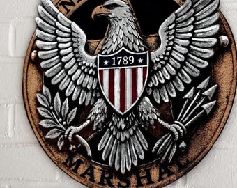 May include: A circular wall plaque featuring a silver-toned eagle with wings spread, clutching an olive branch and arrows. A central shield displays the year 1789 and red and white stripes. The text UNITED STATES MARSHAL is embossed on the border.