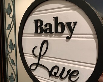 May include: A white round wooden sign with a black border and the words "Baby Love" in black lettering.