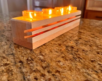 May include: A wooden candle holder with four lit votive candles. The rectangular holder has a natural wood finish with orange accents. The candles are in clear glass holders, casting a warm glow. A decorative item for home decor.