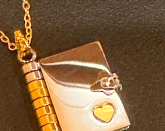 May include: A gold necklace featuring a book-shaped pendant. The pendant has a silver cover, a gold heart, and a gold spine. The chain is fine and gold in colour. The pendant is approximately 2 cm in height.