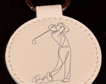 May include: A white leather keychain with a gold-colored ring. The round pendant features a line drawing of a golfer mid-swing, with the word "Golf" in script below. The edges are stitched with white thread.