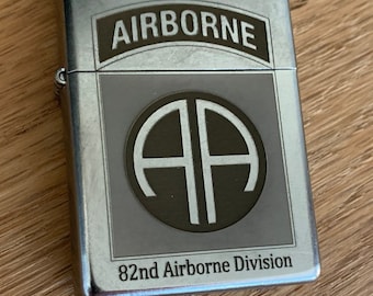 May include: Silver lighter featuring an 82nd Airborne Division emblem. The emblem depicts a circle with the letters "AA" inside and the word "Airborne" above it.