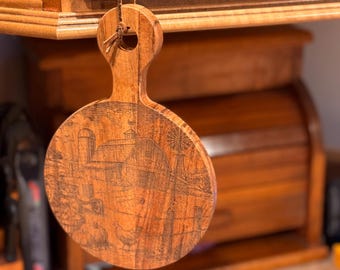 May include: A round wooden cutting board with a handle, featuring a detailed engraved farm scene. The design includes a barn, windmill, tractor, and chickens. The board is suspended by a leather cord, showcasing its rustic charm and functionality for kitchen use.