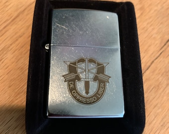 May include: A silver lighter with a black velvet case. The lighter has an engraved image of the Special Forces insignia with the words "De Oppresso Liber".