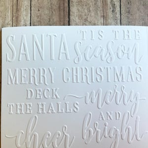 May include: A white rectangular card featuring embossed holiday text that reads: 'Tis the season, Santa, Merry Christmas, Deck the halls, Merry and bright, and Cheer.