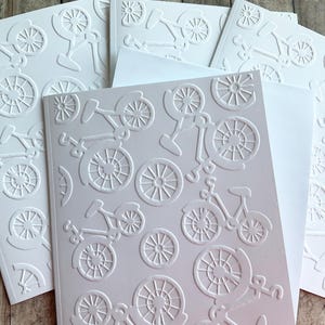 May include: White embossed bicycle-themed greeting cards and envelopes. The cards feature a repeating pattern of bicycles in various sizes. The image shows several cards and envelopes arranged on a wooden surface.