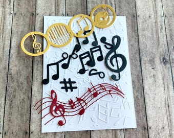 May include: A white card with a musical theme. The card is decorated with black, gold, and red musical notes, a treble clef, and a record player. The background is embossed with musical notes.
