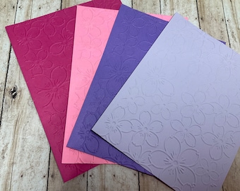 May include: Four sheets of embossed paper with a floral pattern. The sheets are in shades of pink, purple, and lavender.