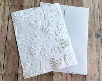 May include: White card with a raised embossed pattern of hearts and swirls. The card is blank and comes with a white envelope.