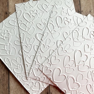 May include: Several white embossed paper sheets with a repeating heart pattern. The hearts are raised, creating a textured surface. The sheets are arranged at an angle, showcasing the design and texture.