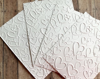 May include: Several white embossed paper sheets with a repeating heart pattern. The hearts are raised, creating a textured surface. The sheets are arranged at an angle, showcasing the design and texture.