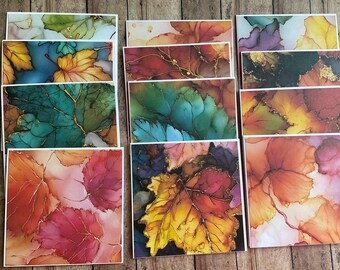May include: A collection of rectangular greeting cards featuring alcohol ink illustrations of autumn leaves in vibrant shades of red, orange, yellow, green, and purple, accented with delicate gold metallic veins throughout the leaf patterns.