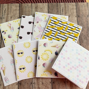 May include: A selection of greeting cards with summer-themed designs. The cards feature illustrations of sunglasses, ice cream cones, bananas, and suns. The colour palette includes yellow, pink, and white, with black accents. The cards are arranged on a wooden surface.