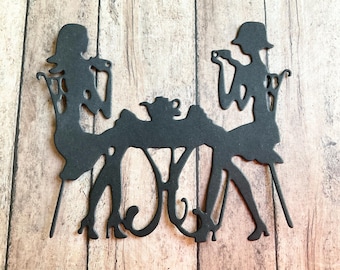 May include: Black silhouette of two women seated at a table, engaged in a tea party. The intricate design includes chairs, a teapot, and delicate details, creating a vintage aesthetic. The figures are wearing hats and dresses.