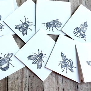 May include: A collection of white square cards, each featuring a detailed, dark gray illustration of a bee. The bees are depicted in various poses, with wings spread and bodies detailed. The cards are arranged on a wooden surface.
