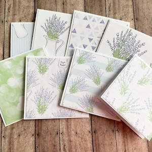 May include: A selection of greeting cards with lavender motifs. Some cards depict lavender sprigs in a white jug, while others display repeating patterns of lavender and geometric shapes. The colour scheme includes shades of purple, green, and white.