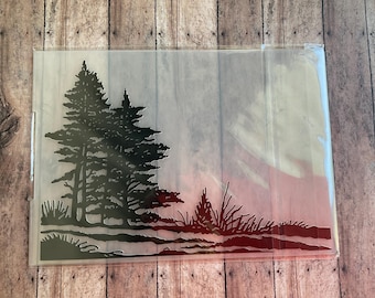 May include: A rectangular, clear plastic stencil sheet featuring a detailed cutout design of a dense forest scene. The design includes tall, layered evergreen trees on the left side, with smaller shrubs and grasses along the bottom edge.