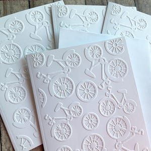 May include: White embossed bicycle cards with matching envelopes. The cards feature a repeating pattern of bicycles and bicycle wheels. The cards are arranged on a wooden surface.