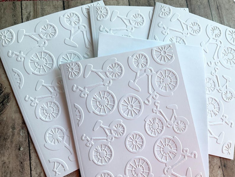 May include: White embossed bicycle cards with matching envelopes. The cards feature a repeating pattern of bicycles and bicycle wheels. The cards are arranged on a wooden surface.