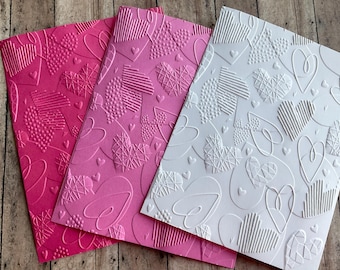 May include: Three embossed paper sheets with a heart pattern. The sheets are pink, light pink, and white.
