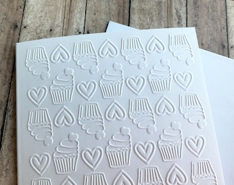 May include: White cardstock with an embossed pattern of cupcakes and hearts. The design features alternating rows of cupcakes with heart-shaped sprinkles and heart shapes. The card is on a wooden surface.