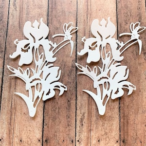 May include: Two white paper cutouts of iris flowers with delicate details. The flowers are on a wood background.