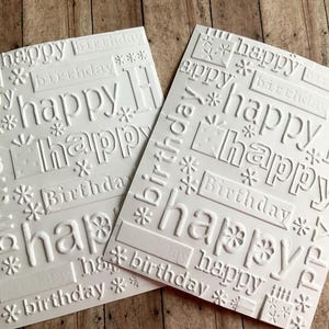 Może przedstawiać: Dwie białe wytłaczane kartki urodzinowe ze słowami "happy" i "birthday" w różnych czcionkach i rozmiarach. Kartki mają teksturowany, wypukły wzór z elementami dekoracyjnymi, takimi jak gwiazdki i pudełka z prezentami. Kartki leżą na drewnianej powierzchni.