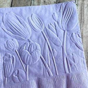 May include: A lavender napkin with an embossed floral design featuring tulips. The embossed tulips are in various stages of bloom, with detailed petals and stems. The napkin has a textured border.