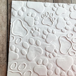 Könnte beinhalten: Weiße Papierserviette mit einem geprägten Muster aus Pfotenabdrücken. Die Pfotenabdrücke sind in verschiedenen Größen und Ausrichtungen.
