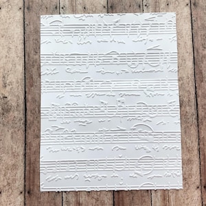 May include: A white rectangular card with an embossed musical staff design. The card features raised lines and symbols representing musical notes and rests. The background is a wooden surface.