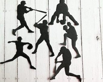 May include: Black silhouette cutouts of baseball players in various poses, including batting, pitching, and catching. The cutouts are on a white wooden background.