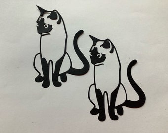 May include: Two black paper cutouts of Siamese cats sitting with their tails curled behind them.