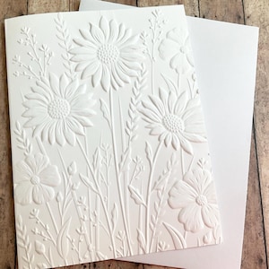 May include: A white embossed greeting card featuring a floral design with daisies and other wildflowers. The card is paired with a blank white envelope, perfect for sending a personalized message. The card is made of thick, textured paper.