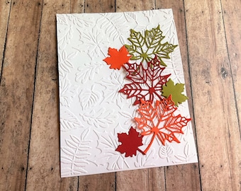 May include: A white card with an embossed leaf pattern, adorned with colorful die-cut maple leaves. The leaves are in shades of red, orange, and green, creating a fall-themed design. The card is set against a wooden background.