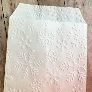 以下が含まれることがあります： 表面全体に雪の結晶のエンボス加工が施された白い紙袋。袋は長方形で上端が折り返されており、さまざまな形の雪の結晶が繰り返されるデザインで、浮き上がった質感のある仕上がりになっています。