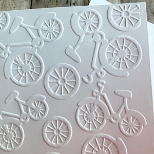 May include: White embossed card with a repeating bicycle pattern. The design features bicycles and bicycle wheels in a raised, textured relief. The card is blank and ready for a personal message.