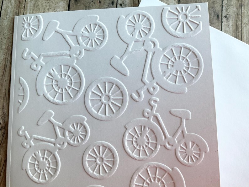 May include: White embossed card with a repeating bicycle pattern. The design features bicycles and bicycle wheels in a raised, textured relief. The card is blank and ready for a personal message.
