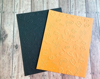 May include: Two rectangular sheets of cardstock, one black and one orange, featuring an embossed pattern of Halloween icons including ghosts, pumpkins, spider webs, bats, skulls, bones, and speech bubbles containing the text BOO!.